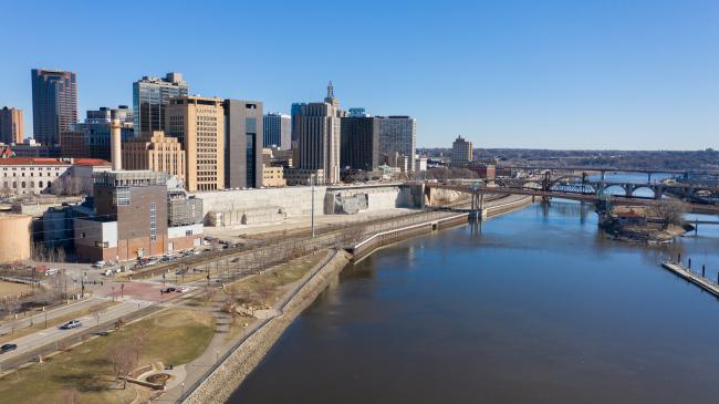 The Mississippi River runs alongside downtown St. Paul, where tall buildings sit on a steep bluff above the river and road.